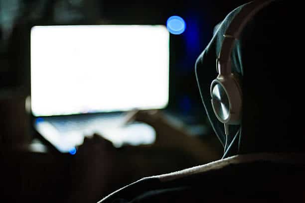 Man hacker in hood and headphones working on laptop in the dark
